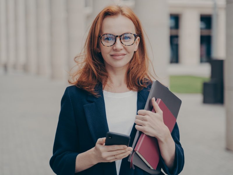 Confident beautiful red-haired female business consultant holding modern smartphone and laptop