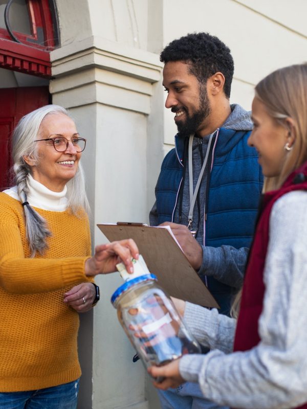 Young door to door fundraisers talking to senior woman and collecting money for charity in street
