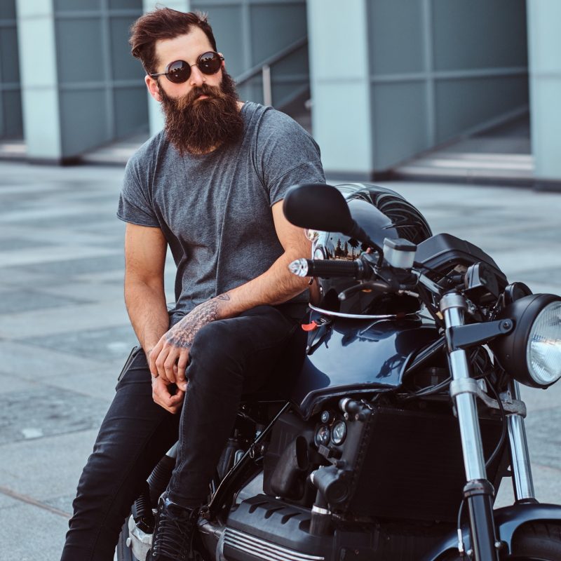 Brutal bearded male in a gray t-shirt and black pants sitting on his custom-made retro motorcycle