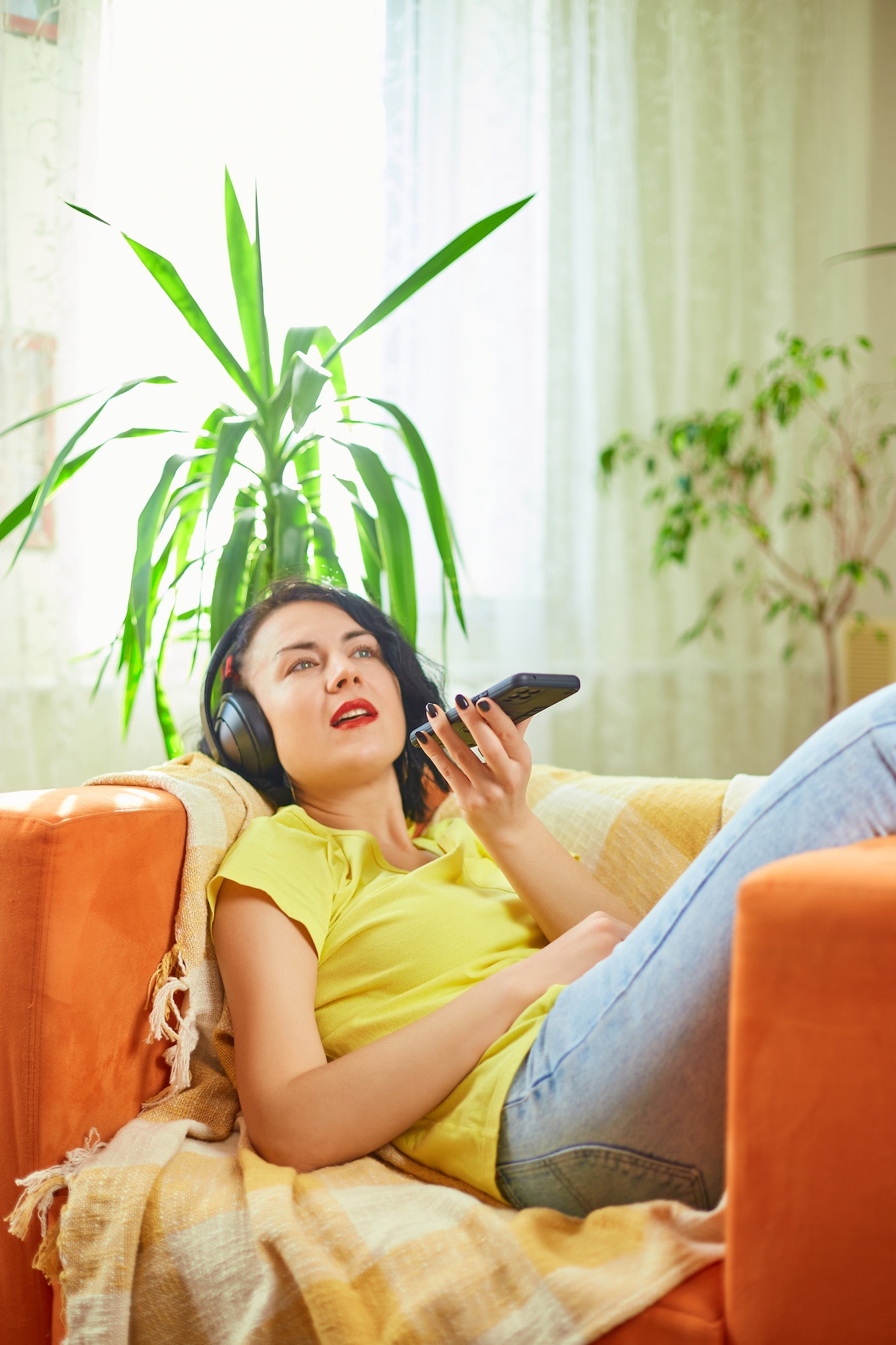 Woman in hedphones, lying on sofa, use virtual commands assistant on smartphone