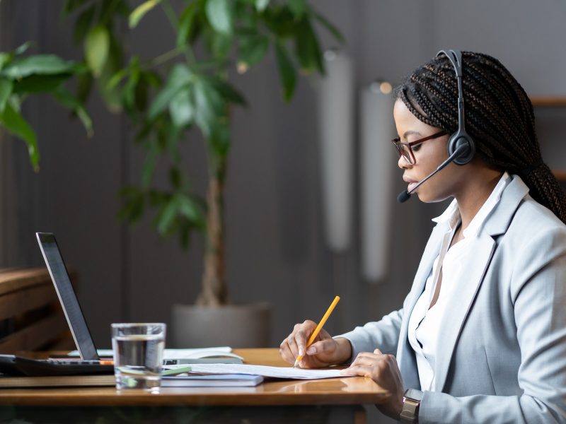 Focused african female home-based virtual assistant in headset making notes during online meeting