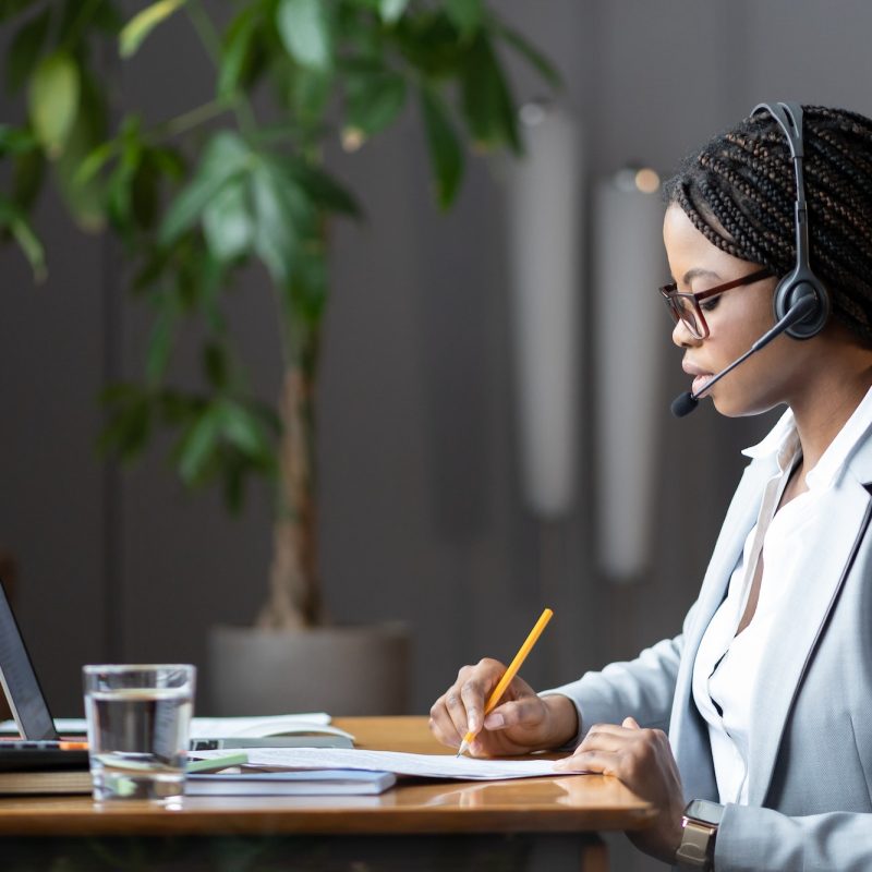 Focused african female home-based virtual assistant in headset making notes during online meeting