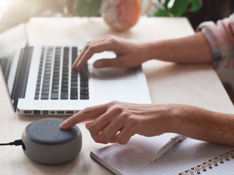 Woman sitting at desk using laptop computer and pressing button on virtual assistant smart speaker