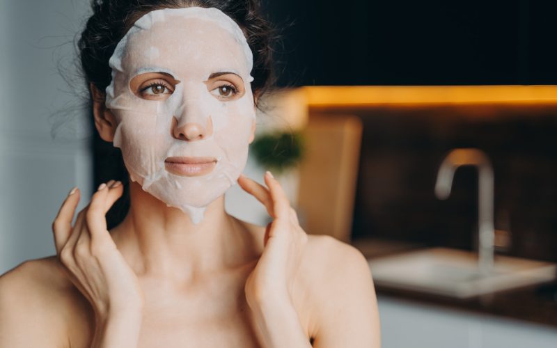 Female with naked shoulders uses rejuvenation facial sheet mask. Skin treatment at home, skincare